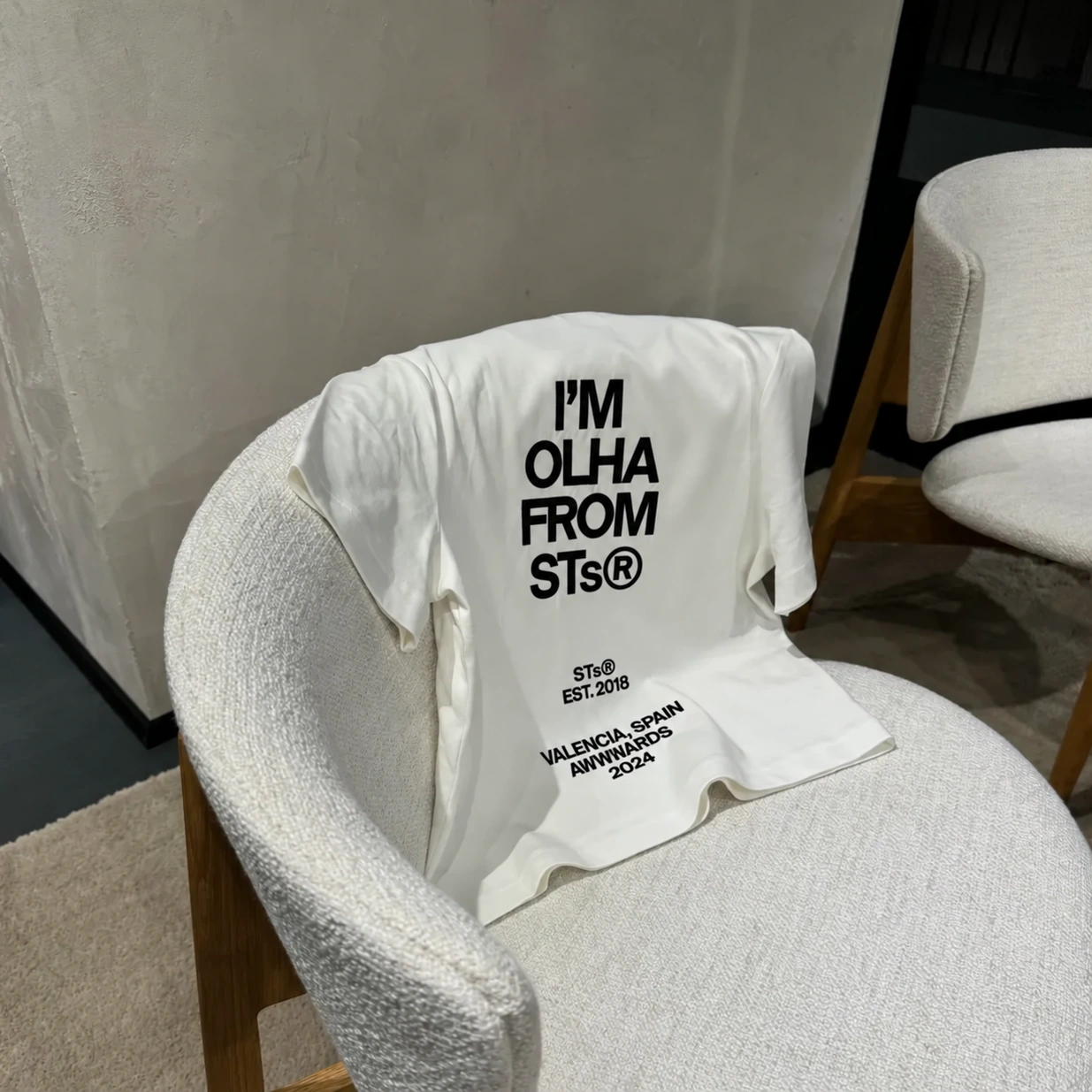 White t-shirt with "I'M OLHA FROM STs®" text draped over a textured white chair.